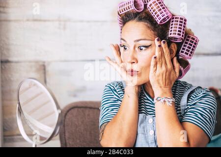Expresión hermosa y divertida mujer caucásica adulta preparándose en casa frente al espejo con maquillaje en la cara - comprobación de arrugas y obtener concepto más viejo para los jóvenes - rizadores sobre el cabello Foto de stock