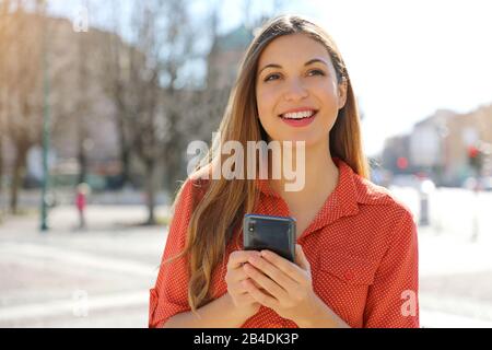 Mujer informal segura sosteniendo el teléfono móvil mirando al lado en la calle de la ciudad