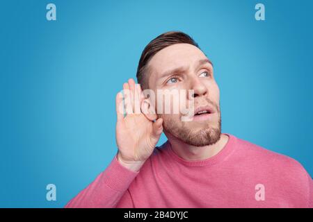 Joven hombre barbudo en el pulóver rosa manteniendo la mano junto al oído derecho mientras escuchas o tratando de oír un sonido suave Foto de stock