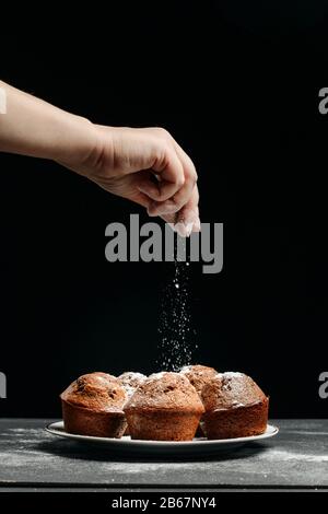 La mano femenina espolvorea magdalenas recién horneadas con azúcar glaseado. Cupcaessalpicado con azúcar glaseado Foto de stock