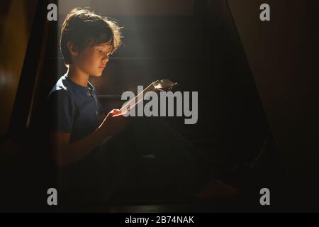 Un niño se sienta en una escalera oscura en un rayo de luz brillante de lectura