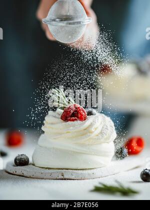 Mujer espolvorea azúcar glaseado a través de un tamiz en mini pastel Pavlova decorado con bayas frescas y romero. Composición vertical. Foto de stock