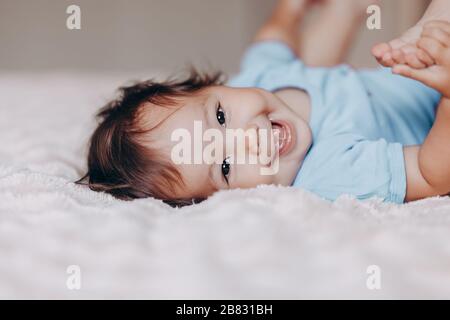 lindo riendo niña de un año acostada en la cama y mirando la cámara tocar sus pies