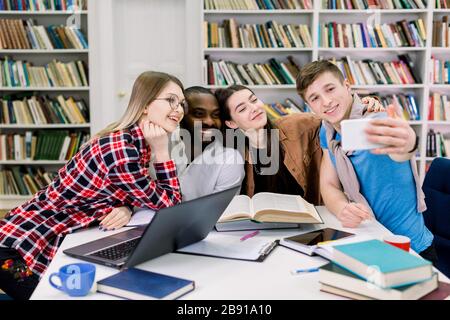Los estudiantes felices sonriendo multiétnicos están estudiando en la biblioteca, divirtiéndose durante el descanso y haciendo fotos selfie. Los jóvenes están pasando tiempo juntos