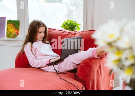 La niña preadolescente disfruta de su hogar en un cómodo pijama y un cómodo sofá-silla. Educación remota, protección antivirus, distanciamiento social y concepto de bloqueo. Flores en la parte delantera Foto de stock