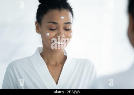 La mujer africana aplicó crema facial haciendo procedimientos de cuidado de la piel en el hogar Foto de stock