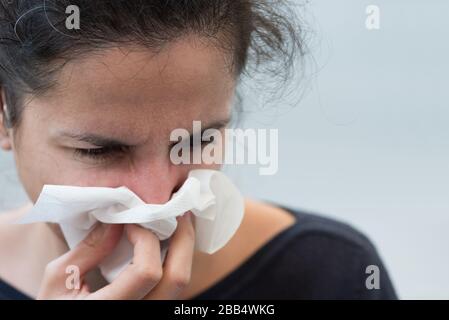 Mujer soplando su nariz en el sofá contra fondo gris. Enfoque selectivo. Gripe, enfermedad, concepto pandémico Foto de stock