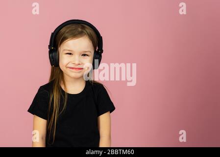 Una niña pequeña con auriculares inalámbricos negros grandes sobre un fondo  rosa pastel mira la cámara. El concepto de escuchar y disfrutar de la  música. Colocar para texto, espacio de copia Fotografía de stock - Alamy