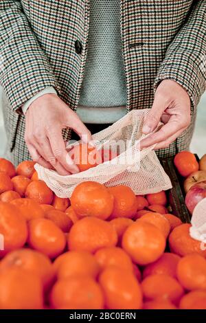 primer plano de un hombre que compra en un greengrocer poniendo algunas naranjas mandarinas en una bolsa de malla textil reutilizable, como medida para reducir la contaminación del plástico Foto de stock