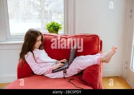 La niña preadolescente disfruta homeschooling en un cómodo pijama y un cómodo sofá-silla. Educación remota, protección antivirus, distanciamiento social y concepto de bloqueo Foto de stock