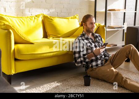 Foto del joven hombre guapo llevando camisa a cuadros sosteniendo el celular y sentado en el piso del apartamento