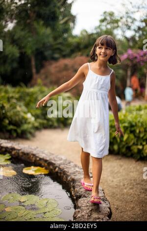 Sonriente joven divirtiéndose equilibrio sobre el muro de piedra de un estanque de jardín.