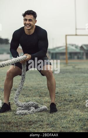 Hombre joven usando cuerdas de entrenamiento para hacer ejercicio al aire libre en un suelo. Atleta tirando de una batalla cuerdas en la lluvia y sonriendo.