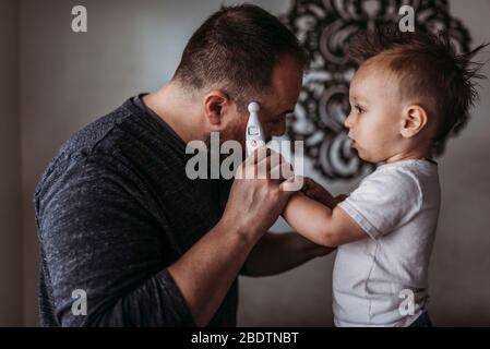 Un niño de un año que toma la temperatura de su padre en casa durante el aislamiento Foto de stock