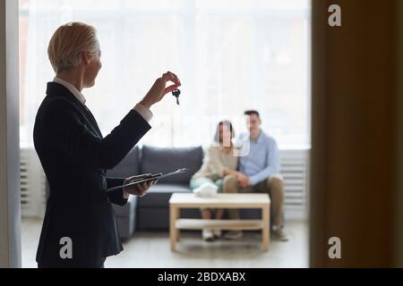 Vista trasera de un realtor en traje negro con llaves de la casa y mostrarlas a una pareja joven que se sienta en el sofá de la habitación