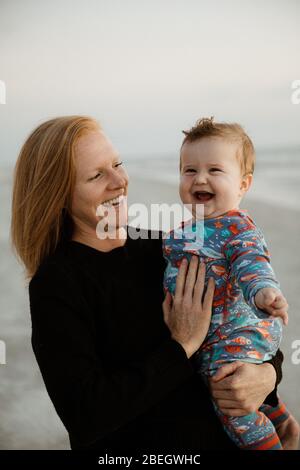mamá soltera con pelo rojo sostiene riendo niño chubby en la playa