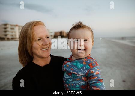 niño gordo con una sonrisa torcida sostenida por una mamá joven de pelo rojo en la playa