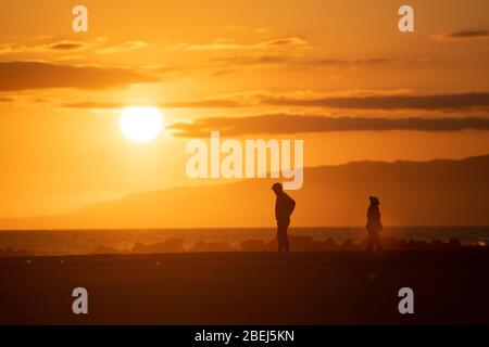 Los Ángeles, EE.UU. 13 de abril de 2020. Dos personas están de pie en la playa de Venice Beach viendo la puesta de sol. Crédito: Maximilian Haupt/dpa/Alamy Live News