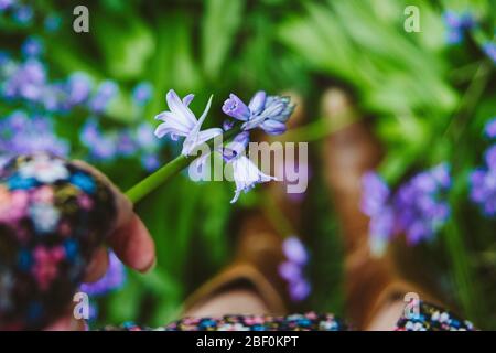 Una niña o mujer escogiendo un bluebell en un día de primavera, usando un vestido floral y botas de vaquero. Céntrese en el bluebell en la mano. Profundidad de la dof