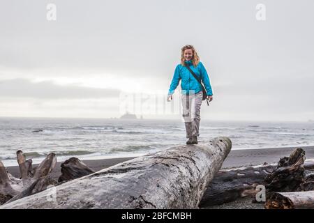 Una mujer caminando sobre un gran tronco de madera a la deriva sobre una playa. Playa Rialto, Parque Nacional Olímpico, Washington, Estados Unidos.