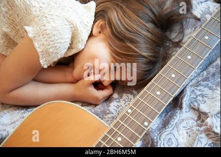 Una niña soñadora acostada en la cama con su guitarra acústica. Ella sonríe con la mano en la cara. Ella bromeaba con su guitarra. El sueño es ser un Foto de stock