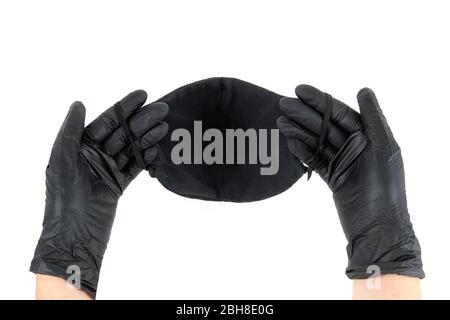 Las manos femeninas en guantes desechables negros sostienen una máscara facial reutilizable negra hecha de algodón. Vista en primera persona, aislada sobre blanco. Foto de stock