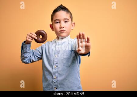 Niño pequeño joven comiendo donut de chocolate malsano sobre fondo amarillo aislado con la mano abierta haciendo stop signo con exp serio y confiado Foto de stock