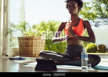 Mujer sana sentada en la meditación del yoga en casa con una tableta digital en frente. Mujer de fitness en sportswear meditando en lotus yoga pose en el interior.