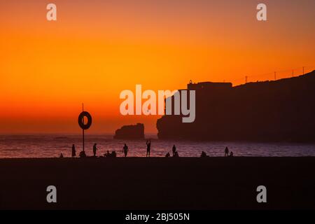 Nazare, Portugal: Increíble puesta de sol naranja sobre el Océano Atlántico y la gente que se relaja en la playa. Faro y fuerte de Sao Miguel visible en la parte alta Foto de stock