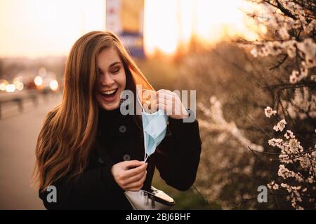 Feliz alegre joven mujer quitando la máscara médica de la cara mientras que está parado en la calle en la ciudad durante la puesta de sol en primavera