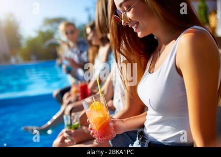 Bonita mujer relajarse con un colorido cóctel de vidrio fresco sentado junto a la piscina el soleado día de verano con amigos. La gente tosta bebidas en Foto de stock