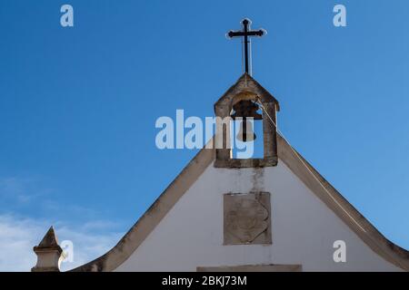 Triángulo de una pared y techo. Fachada blanca y detalles de piedra. Campanario sencillo con una campana y una cruz en la parte superior. Cielo azul brillante. Alvor, Algarve, Portug