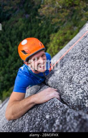 escalador mirando hacia arriba y riendo mientras escalaba en roca con casco Jefe Foto de stock
