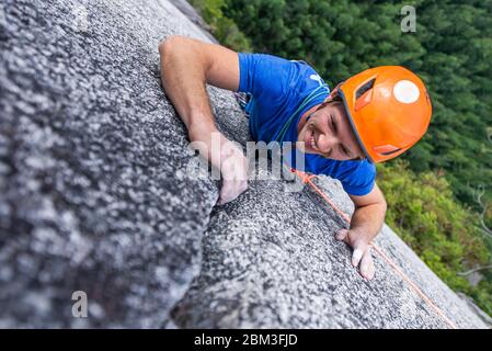 escalador mirando hacia arriba y riendo mientras escalaba en roca con casco Jefe Foto de stock