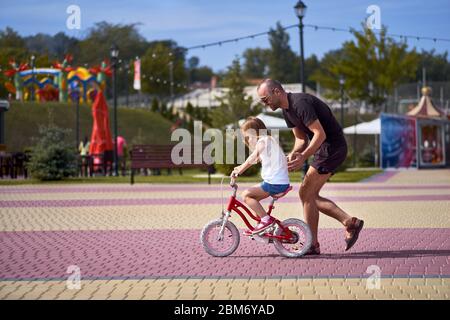 Retrato de un padre feliz y cariñoso enseñando a su pequeña y bonita hija en bicicleta en un parque verde, sonriendo a lo largo de toda la vida