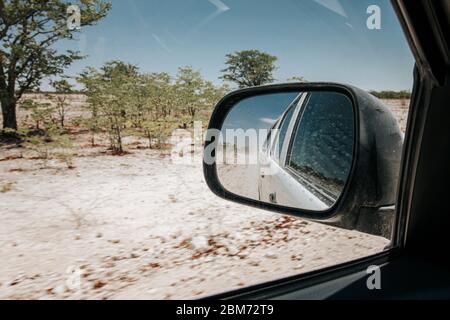 Vista en el espejo retrovisor mientras conduce por el árido paisaje seco en el parque nacional Etosha Namibia. Foto de stock