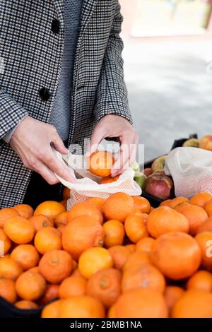primer plano de un hombre que compra en un greengrocer poniendo algunas naranjas mandarinas en una bolsa de malla textil reutilizable, como medida para reducir la contaminación del plástico Foto de stock