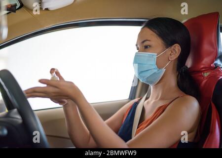 Mujer asiática joven con máscara para la protección contra el brote del virus de la corona rociando alcohol a mano dentro del coche Foto de stock