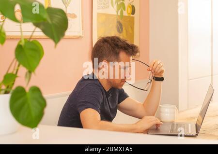 Retrato de un hombre trabajando desde casa en un ordenador portátil durante el bloqueo del coronavirus. Concentrarse mientras muerde los vasos. Formato horizontal. Foto de stock