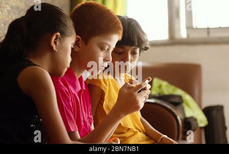 Tres niños multiétnicos o hermanos ocupados en juegos móviles en casa - concepto de niños móviles de juego de vídeo adicción, utilizando la tecnología Foto de stock