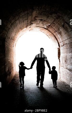 Padre caminando con sus dos pequeños hijos hermanos, chico y chica, hacia la luz al final de un largo túnel oscuro Foto de stock