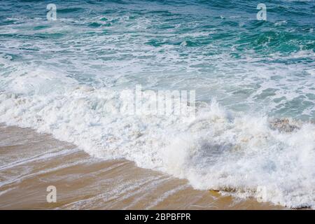 Agua turquesa del océano con pequeñas olas de espuma blanca que rompen suavemente en la playa de arena dorada