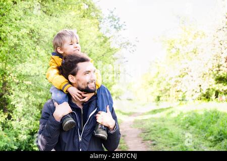 Feliz paseo familiar al aire libre. Padre montando a su hijo pequeño sobre sus hombros en un parque. Día de los padres y familia divertido concepto