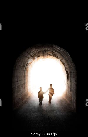 Dos pequeños hermanos, hijos, niño y niña, caminando juntos y caminando hacia la luz al final de un largo túnel oscuro Foto de stock
