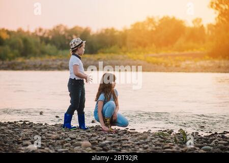 Rubio chico en sombrero y chica jugando en la orilla del río. Botas de goma para niños. Actividades de primavera