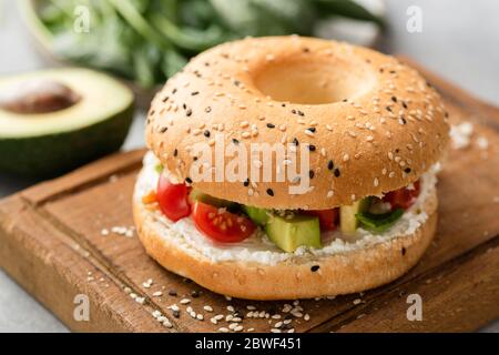Sándwich de bagel con queso crema, aguacate y tomate en tabla de servir de madera, Vista de primer plano. Hamburguesa vegetariana con bagel de sésamo