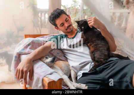 Un joven sentado en un sillón en el jardín, acaricia la cabeza de un gato negro