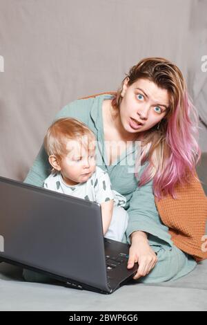 Quédese en casa, mamá trabaja remotamente en un portátil, cuidando a su hijo. Una joven madre que está de baja por maternidad está tratando de trabajar como una freelancer con un bab Foto de stock