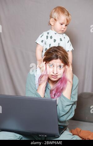 Quédese en casa, mamá trabaja remotamente en un portátil, cuidando a su hijo. Una joven madre que está de baja por maternidad está tratando de trabajar como una freelancer con un bab Foto de stock