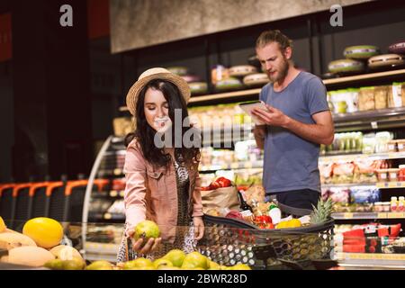 Joven hombre serio pensativamente leyendo lista de compras mientras sonriente chica en sombrero elegir frutas con carrito lleno de productos en el supermercado moderno Foto de stock